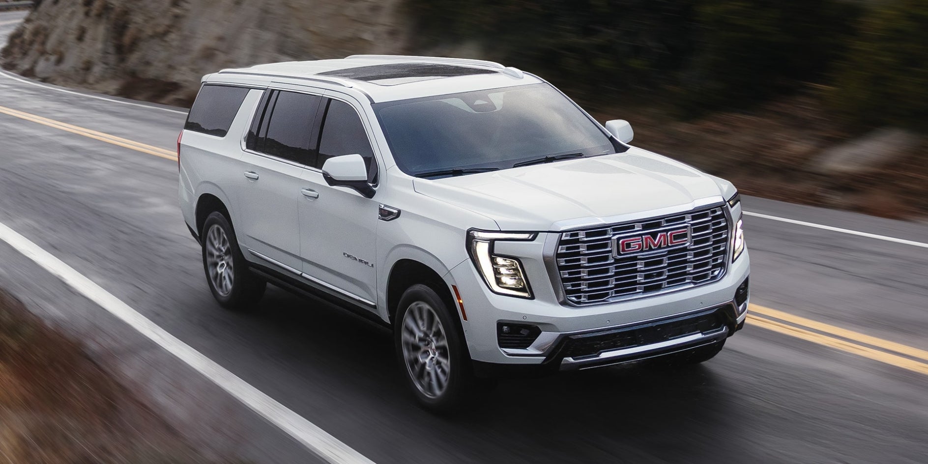 A white GMC Yukon driving down the road near a cliffside.