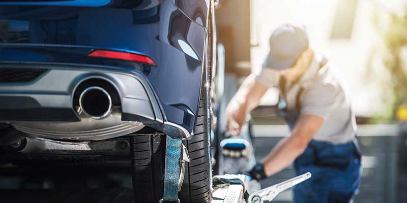 A blurred person in a cap and gloves secures a blue car onto a tow truck with straps.