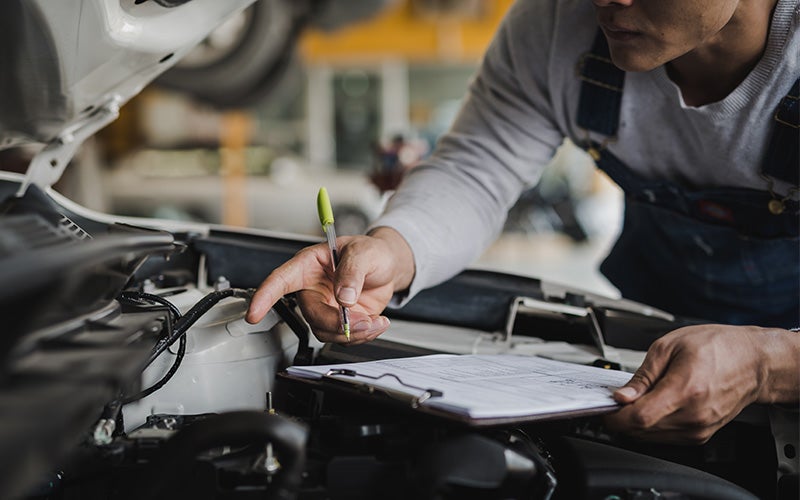 Mechanic in overalls inspecting a car engine and holding a pen and clipboard to record findings.